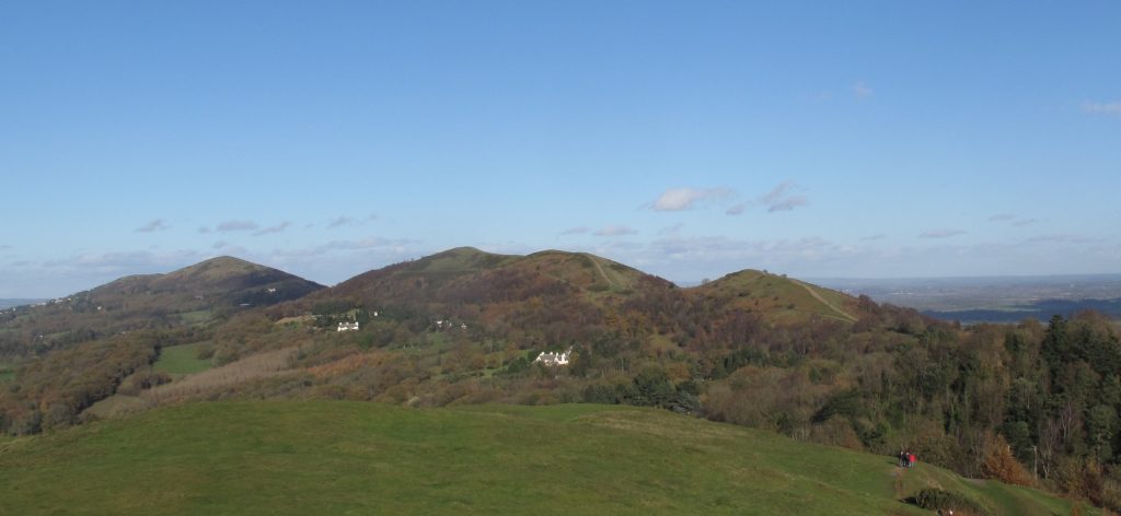a line of hills in the distance, with a mix of tree cover and grass a few paths are visible. There is blue sky and a few clouds. In the foreground is a grassy area of the hill the photographer is standing on.