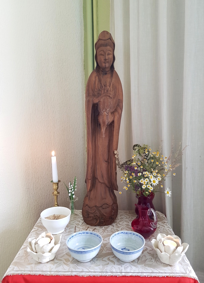 A wooden statue, of Quan Yin, a female Buddhist Saint, wearing flowing robes and holding an upturned vase on a small alter. The statue is about one and a half feet tall. There is a small red vase on the right, full of wildflowers, a candle stick with a lit cnadle on the left and two small offering bowls filled with water in front.