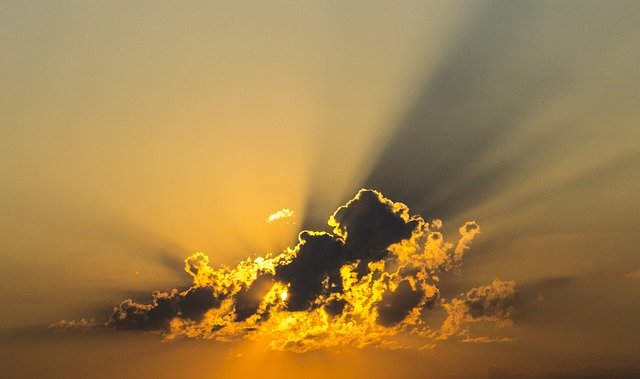 the sun is behind a few small clouds, the sky is yellow orange, and streaks of shadow cross the sky from the clouds. The clouds have golden edges