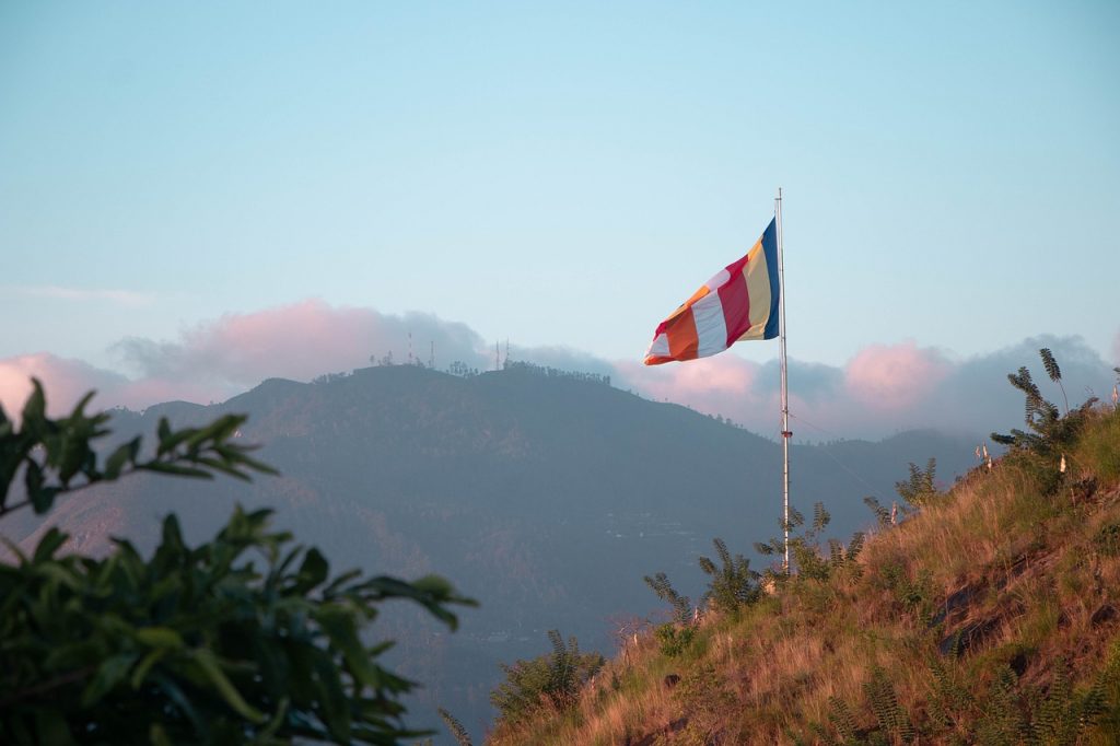 On the side of a hill there is a flag pole, the Buddhist flag waves from the top of the pole. The Buddhist flag is vertical stripes of orange, white, red yellow and blue..